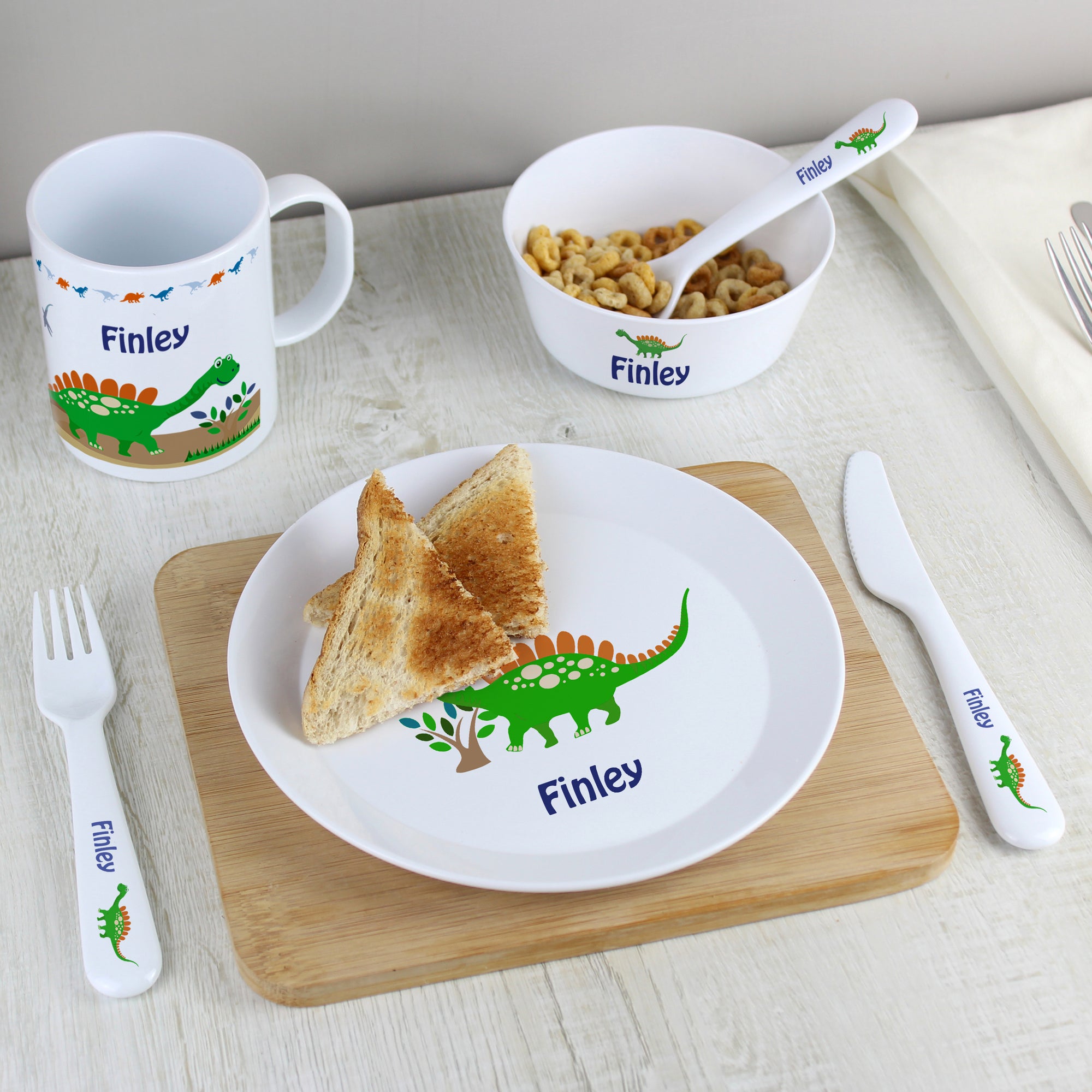 Image of a table layed with a personalised plate, cup, bowl and cutlery set made from white shatter resistance BPA free plastic.  The set features a green dinosaur on each piece and they can be personalised with a name of your choice of up to 12 characters.