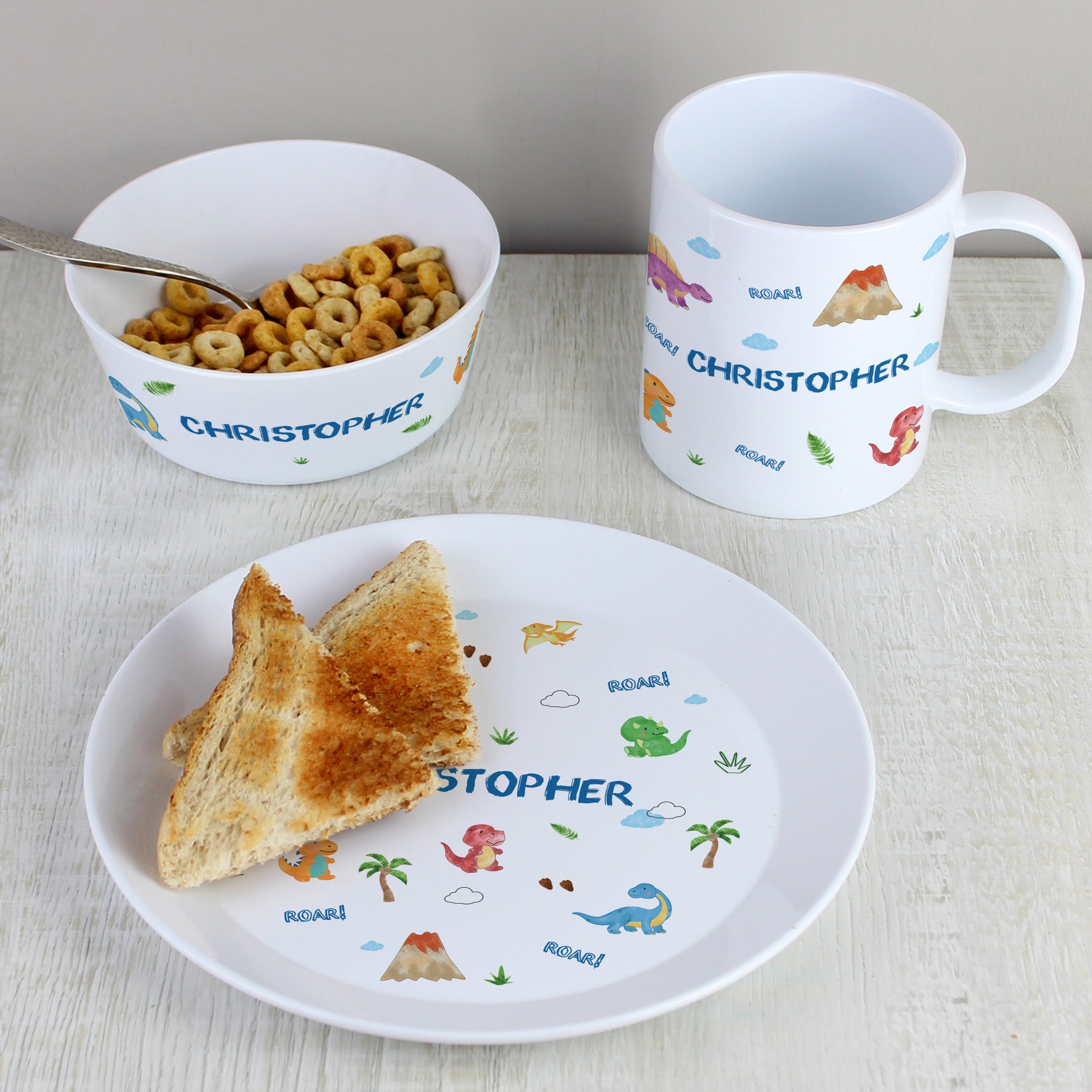 Image of a personalised white plate, cup and bowl made from BPA free plastic.  The set has a dinosaur design on it which is made up of small dinosaur images and palm tree images. The set is designed for children and is shatter proof.