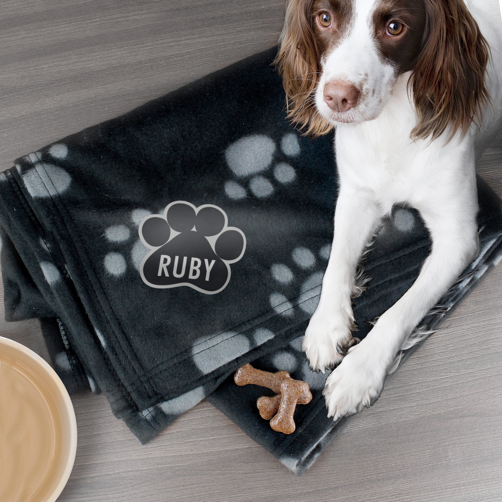 Image of a brown and white cocker spaniel dog sitting on a personalised dog blanket.  The blanket is made from black fleece with a grey pawprint design and the blanket can be personalised with a name of your choice of up to 12 characters which is printed in the corner of the blanket within a paw print image. 