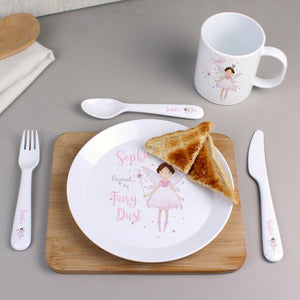 Image of a white plastic BPA free childs personalised plate, cup and cutlery set, laid on a table ready to use. Each piece in the set is made from strong white plastic and has a fairy holding a wand printed on it with the text 'Powered by Fairy Dust'. Each piece in the set can be personalised with a name up to 12 characters in a pink font. 