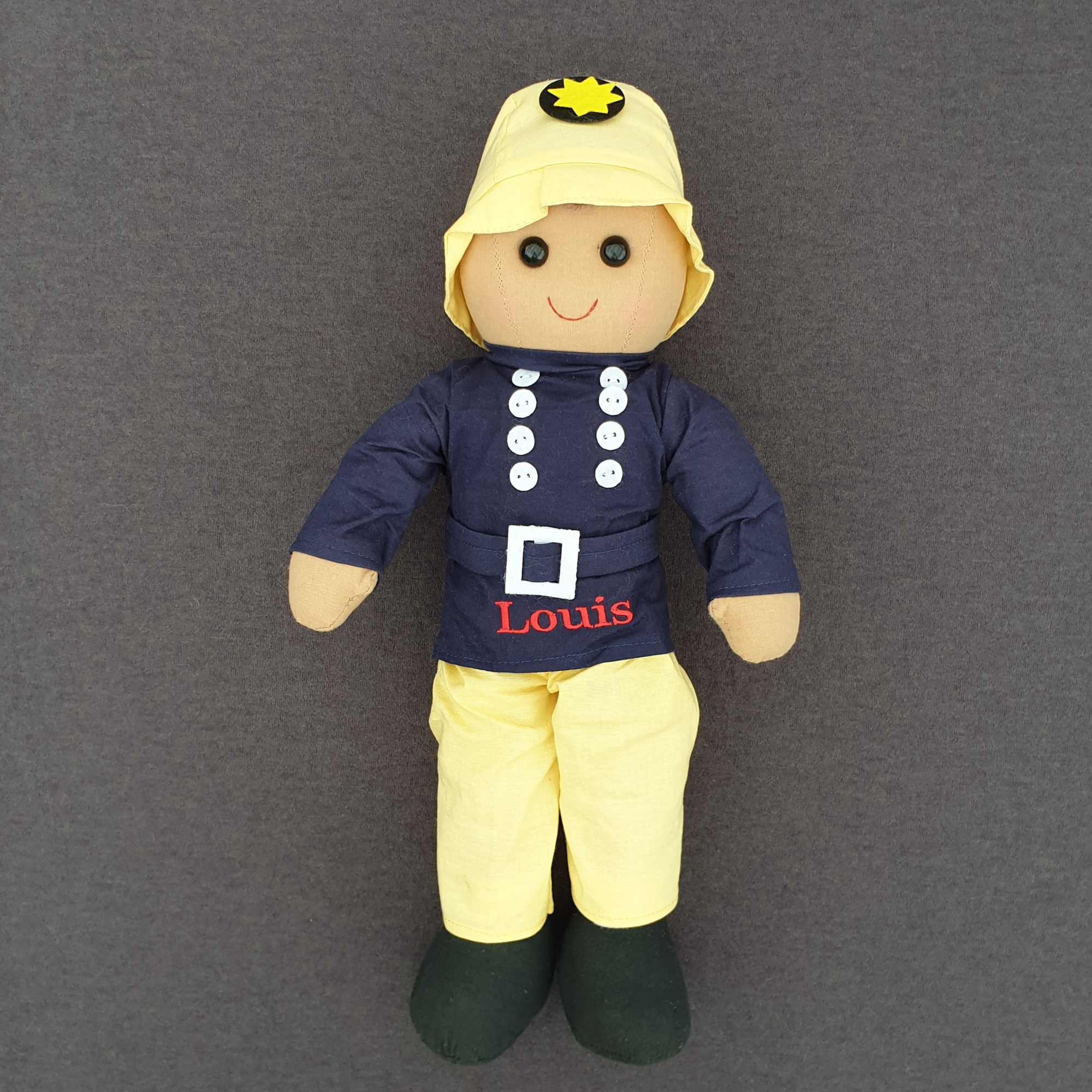 Personalised fireman ragdoll. The doll is wearing yellow trousers and a yellow hat along with a navy blue tunic with a belt and white buttons. The doll is made from cotton and measures approximately 40 cm in height.