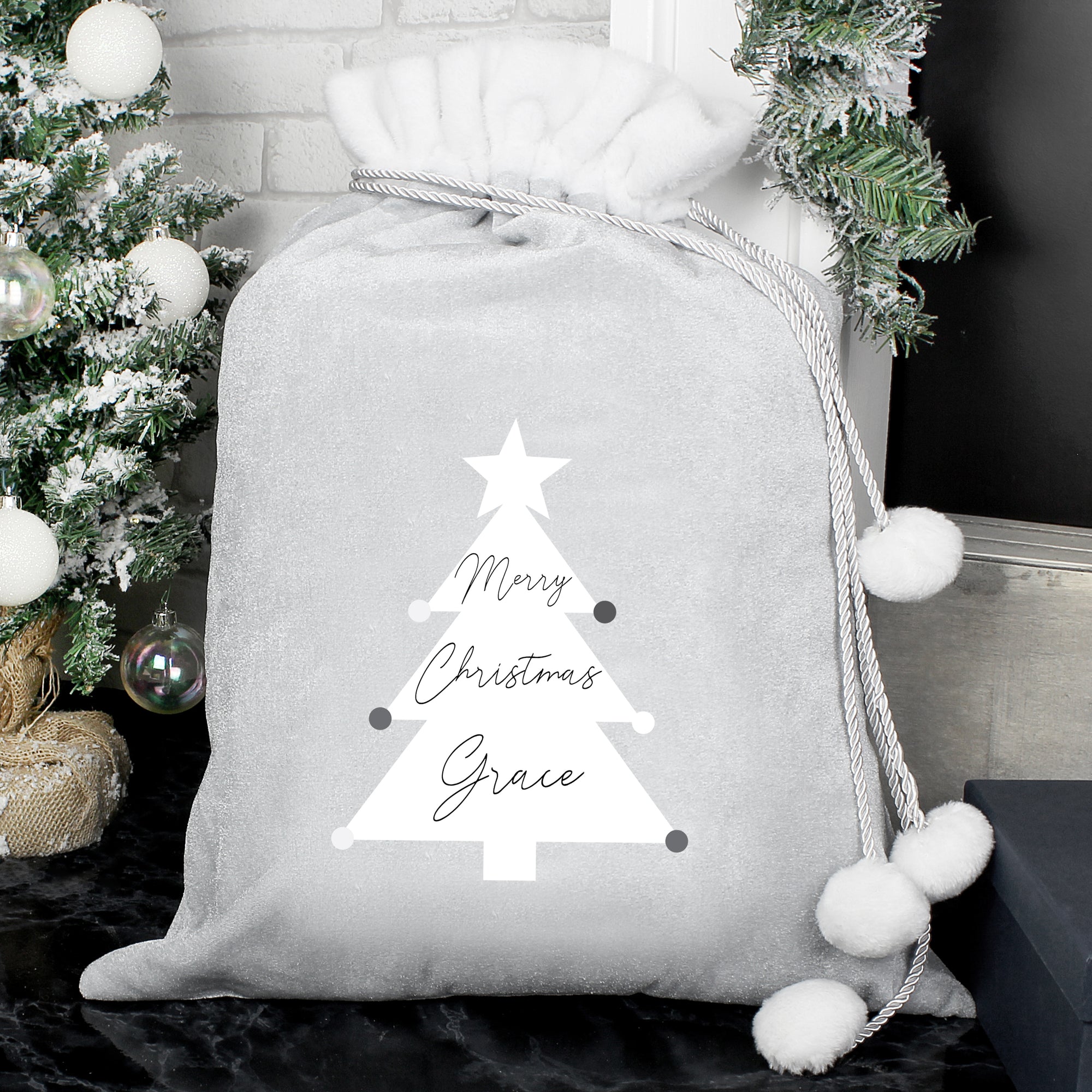 Personalised grey Christmas present sack with a white fur trim and white drawstrings with pom poms on the end to close the sack. The front of the sack features an illustration of a white Christmas tree and the sack can be personalised with a name of your choice up to 12 characters long which will be printed in a black modern cursive font within the image of the tree along with the words 'Merry Christmas'.