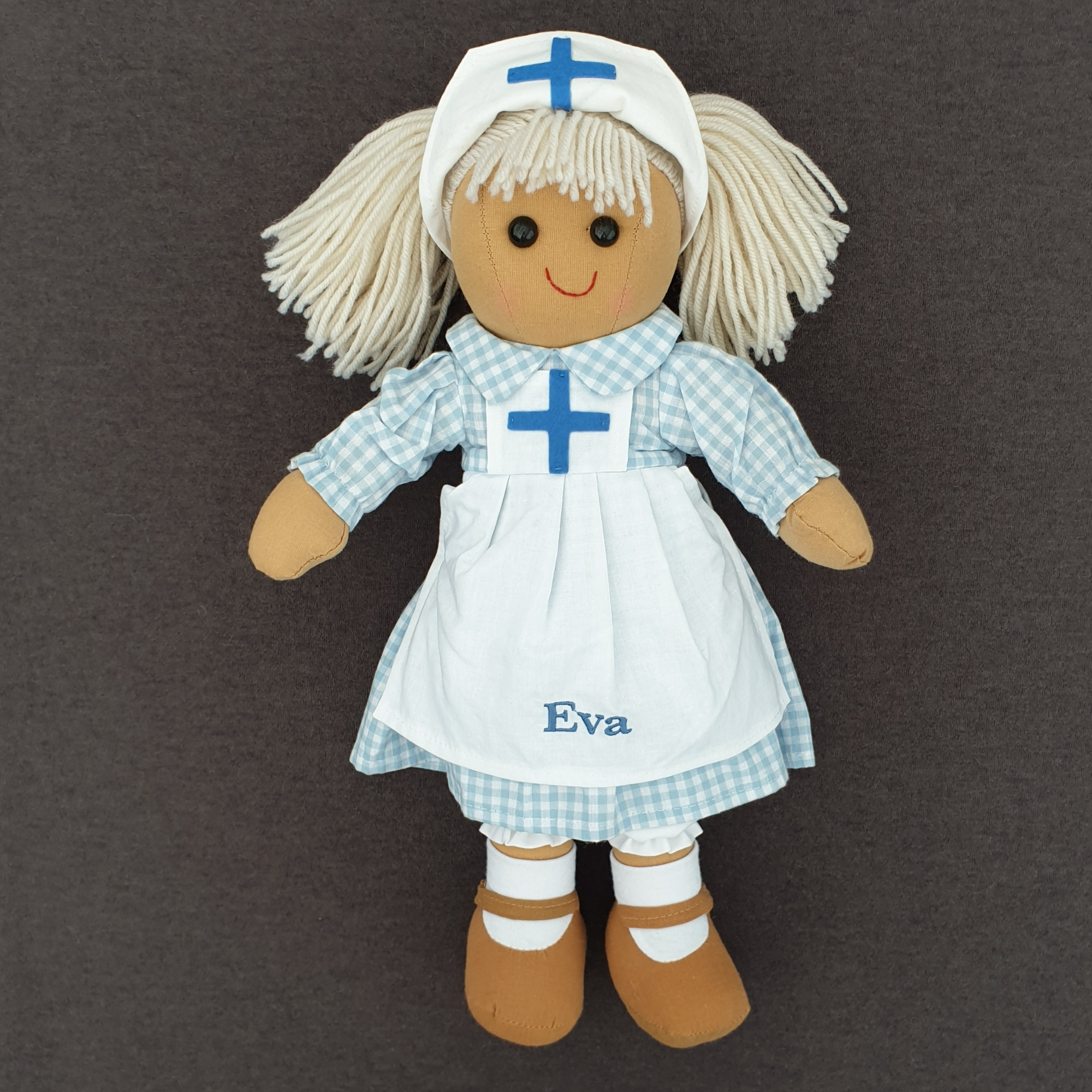 Personalised nurse rag doll. The doll is wearing a blue and white gingham dress with a white apron over the top that can be embroidered with a name of your choice.  She has blonde hair in bunches and a white hat with a blue cross on it.
