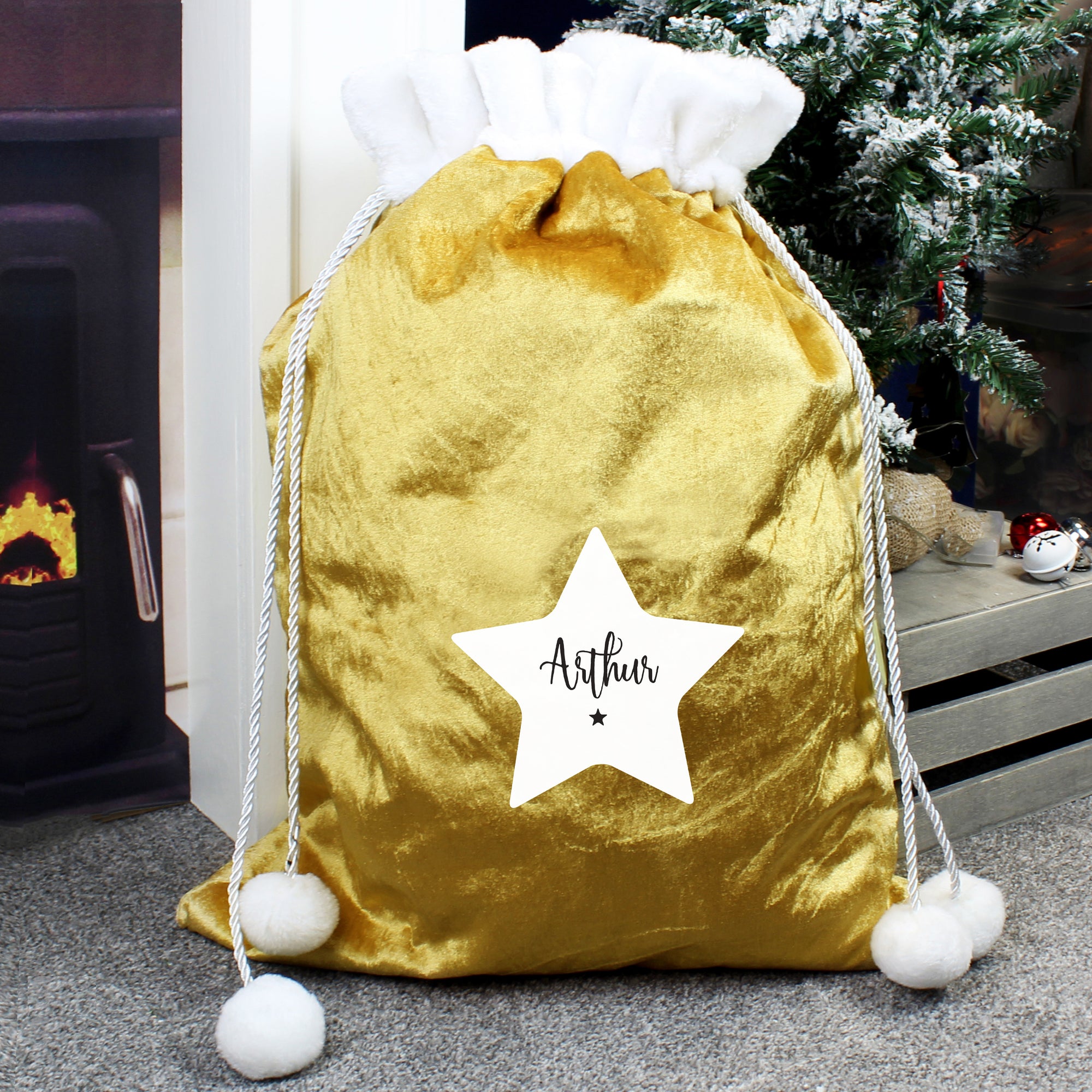 Personalised plush gold Christmas present sack with a white fur collar at the top. The sack can be closed with drawstrings which have white pom poms on the end. The front of the sack features a white star and a name of your choice of up to 12 characters will be printed inside the star in a black modern cursive font.  There is also a small black star printed underneath the name.