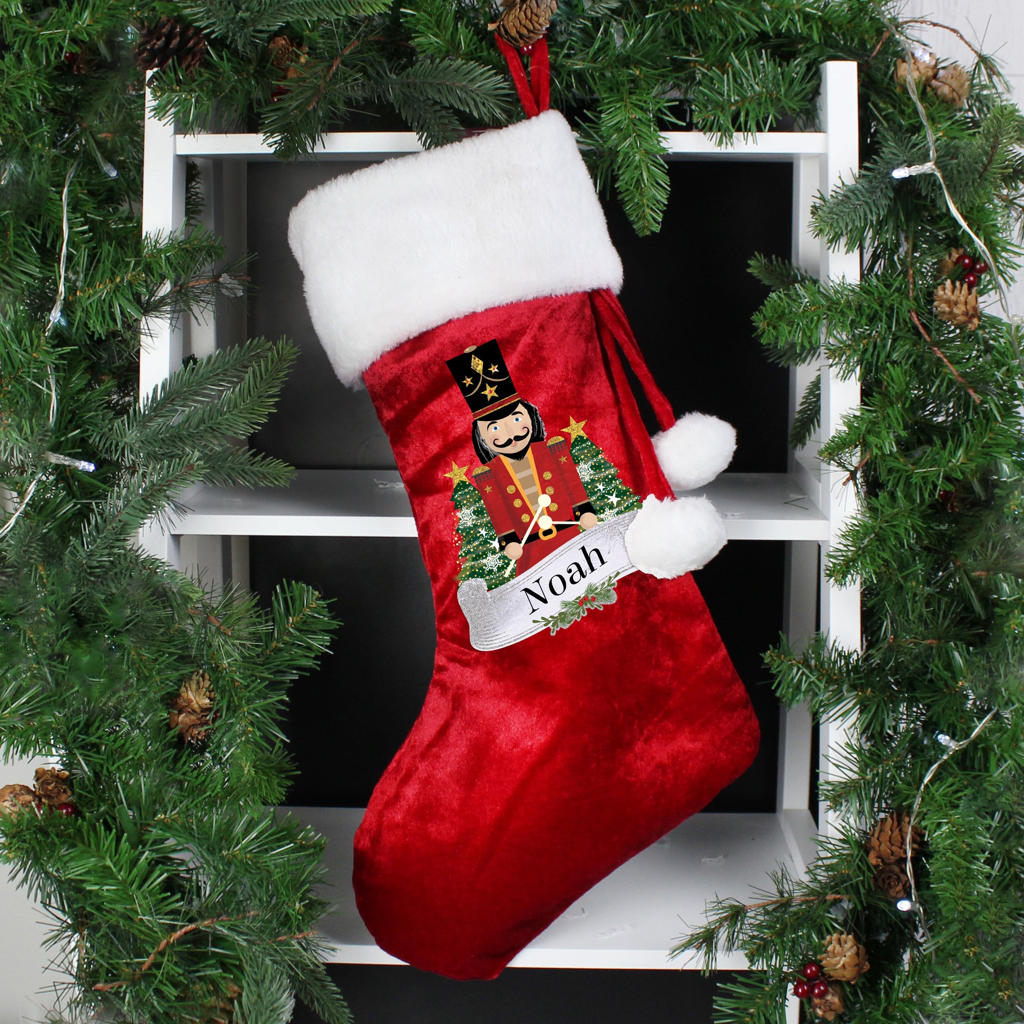 Plush red Christmas stocking with a white fur trim and two white pom poms hanging from the side. The stocking has an illustration of a wooden nutcracker toy on the front and it can be personalised with a name of your choice of up to 12 characters.