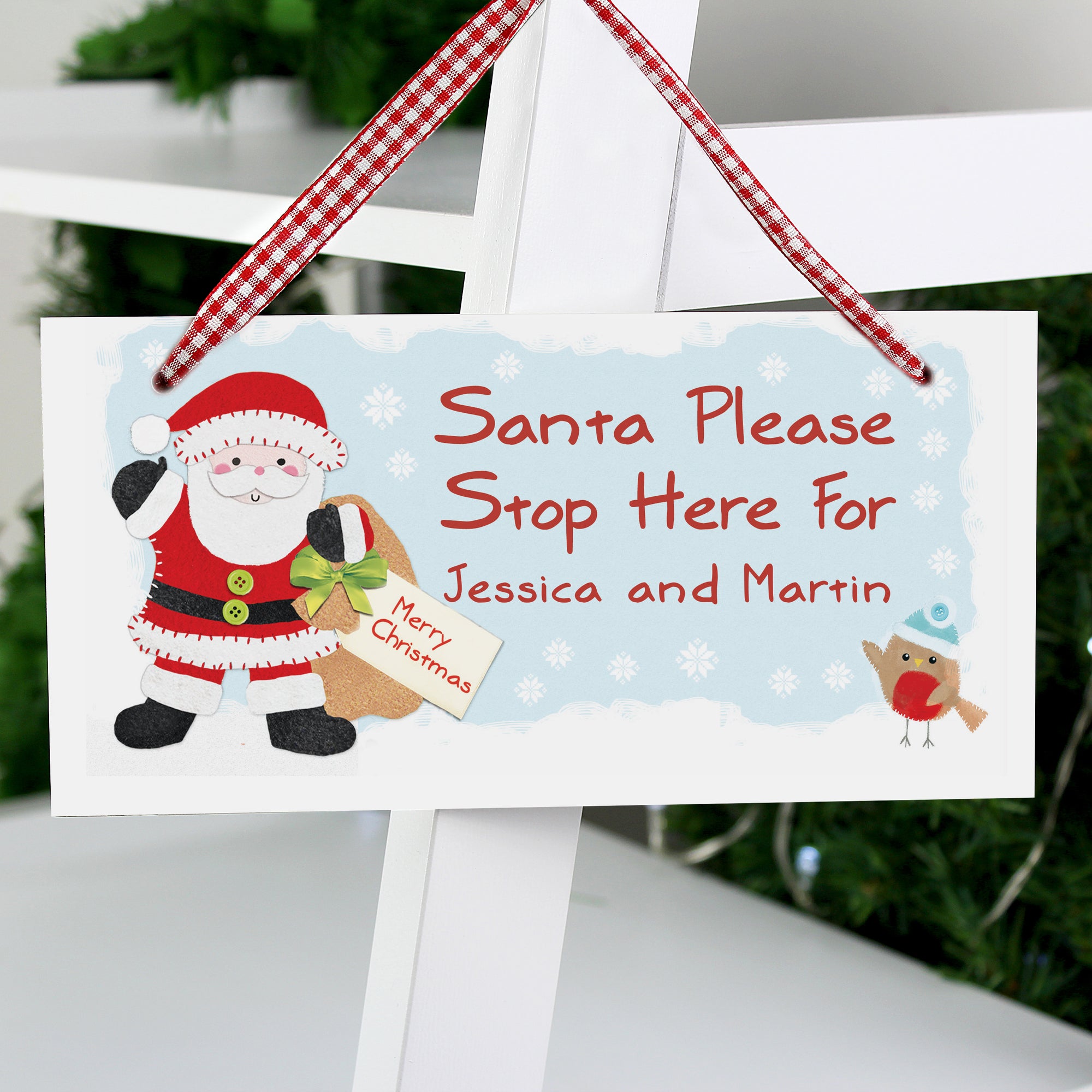Personalised wooden sign printed with a cartoon Santa character holding a Christmas present sack and a robin. The wooden sign features the text 'Santa Please Stop Here For' and then your own names can be added. The sign comes with a ribbon to hang it up and measures approximately 20 cm by 10 cm.