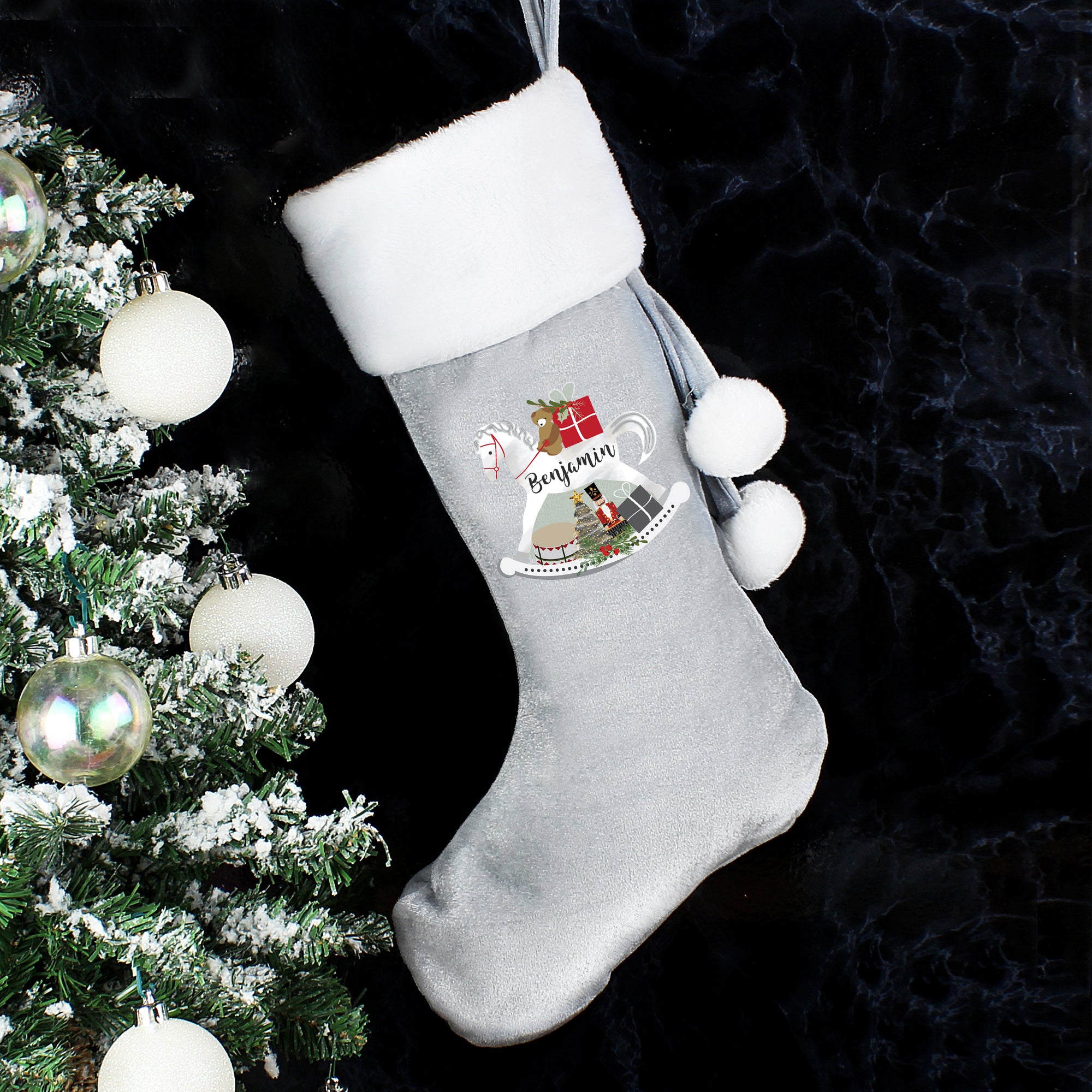Personalised silver velvet Christmas stocking featuring an illustration of a rocking horse on the front which can be personalised with a name of up to 12 characters which will be printed in a modern black cursive font on the body of the rocking horse. The stocking has a white plush cuff and has a loop at the top so it can be hung from a hook.