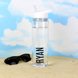 Image of a transparent reuseable water bottle sitting on some sand with a blue sky behind it and a pair of sunglasses lying next to the bottle.  The bottle is made from BPA free transparent hard wearing plastic with a white screw on lid that has a flip up spout which is connected to a straw inside the bottle.  The outside of the bottle can be personalised in a name of  your choice of up to 12 characters which will be printed in a large black uppercase font on the side of the bottle.