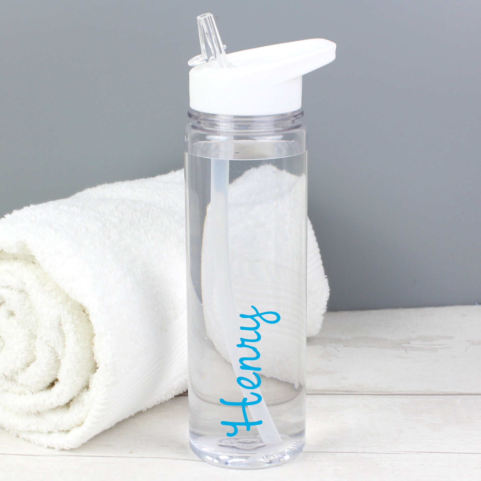 Image of a transparent reuseable water bottle filled with water standing next to a rolled up white towel. The bottle is made from BPA free transparent hard wearing plastic with a white screw on lid that has a flip up spout which is connected to a straw inside the bottle.  The outside of the bottle can be personalised in a name of your choice of up to 12 characters which will be printed in a blue font on the side of the bottle.