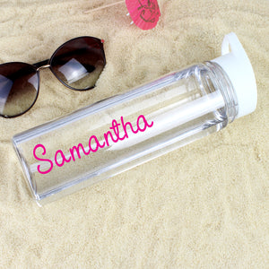 Image of a transparent reuseable water bottle lying on some sand next to a pair of sunglasses and a red cocktail drinks umbrella.  The bottle is made from BPA free transparent hard wearing plastic with a white screw on lid that has a flip up spout which is connected to a straw inside the bottle.  The outside of the bottle can be personalised in a name of  your choice of up to 12 characters which will be printed in a pink font on the side of the bottle.