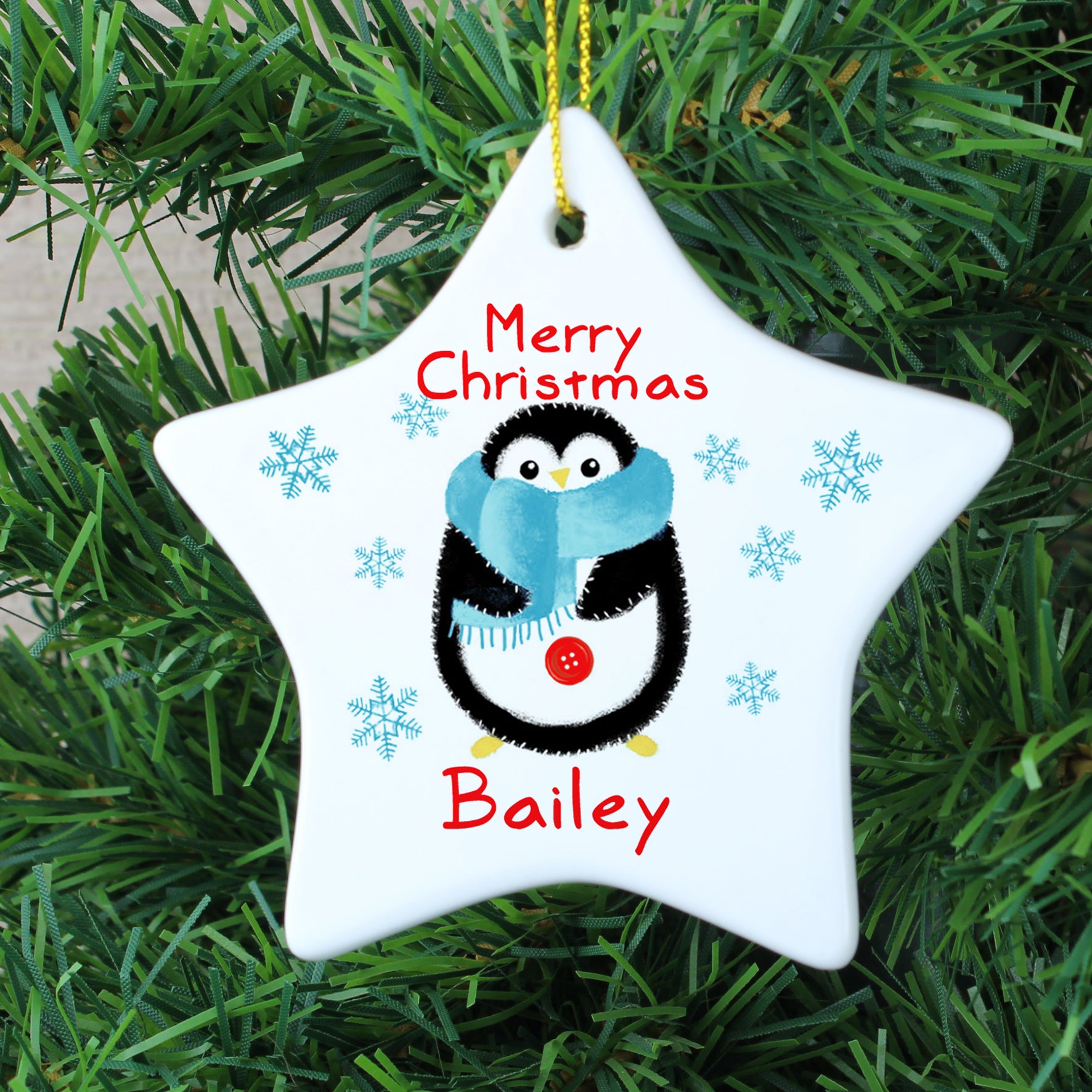 Personalised white ceramic Chrismas star decoration featuring the words 'Merry Christmas' in a red font and an image of a hand drawn penguin wearing a blue scarf. The decoration can be personalised with a name in red which will be printed below the image of the penguin.