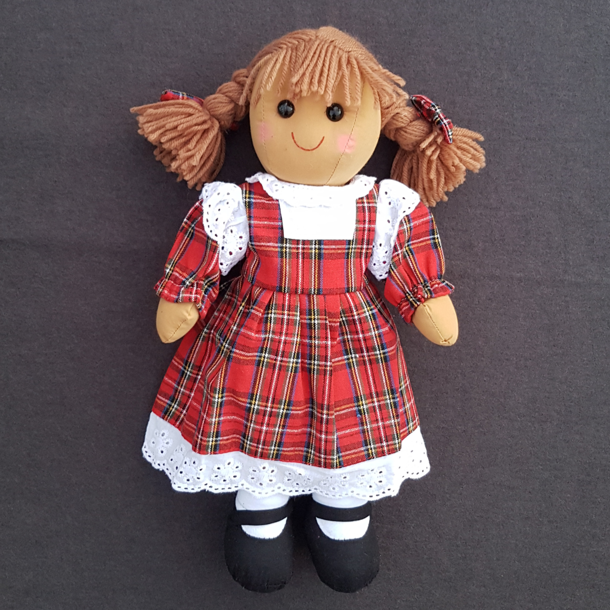 image of a personalised rag doll with brown hair in plaits and wearing a red Scottish tartan dress. The dress has white trim at the top of the arms and around the hem of the dress. Underneath the dress she is wearing white pantaloons and she has black cloth shoes sewn onto her feet. The dolls dress can be personalised with a name of your choice which will be embroidered in white thread.